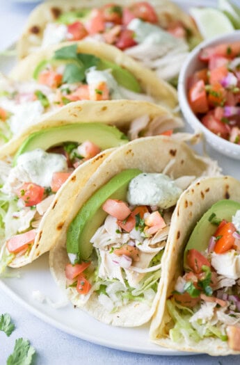 Close-up of tacos with creamy avocado, onion-pepper mixture, cilantro crema, shredded chicken, and lime juice.