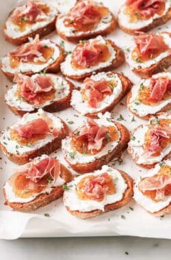 Angled photo of crunchy crostinis with creamy goat cheese, fig jam, and salty prosciutto on top.