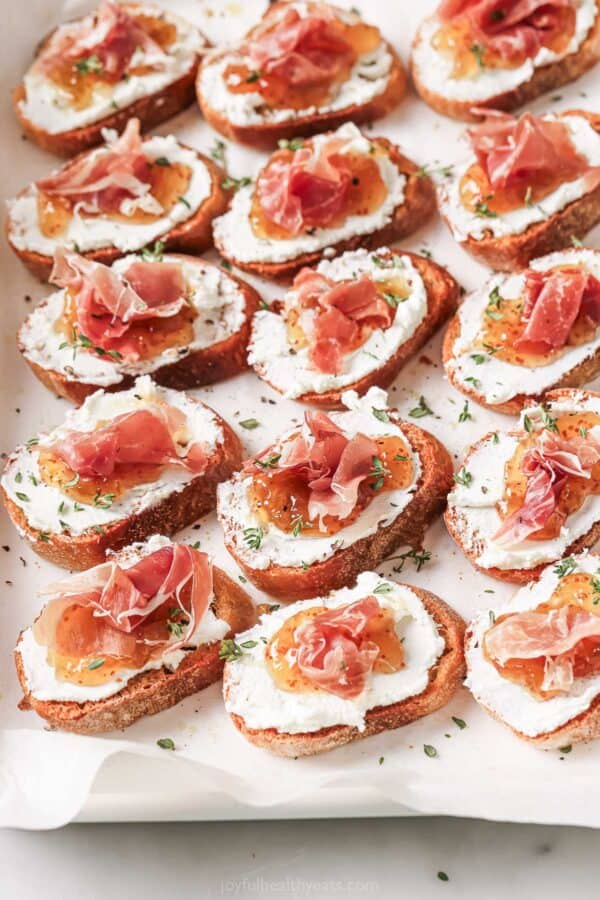 Angled image of crunchy crostinis with creamy goat cheese, fig jam and salty prosciutto on top.