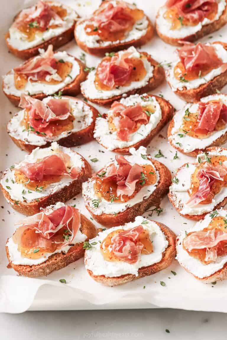 Angled photo of crunchy crostinis with creamy goat cheese, fig jam, and salty prosciutto on top.