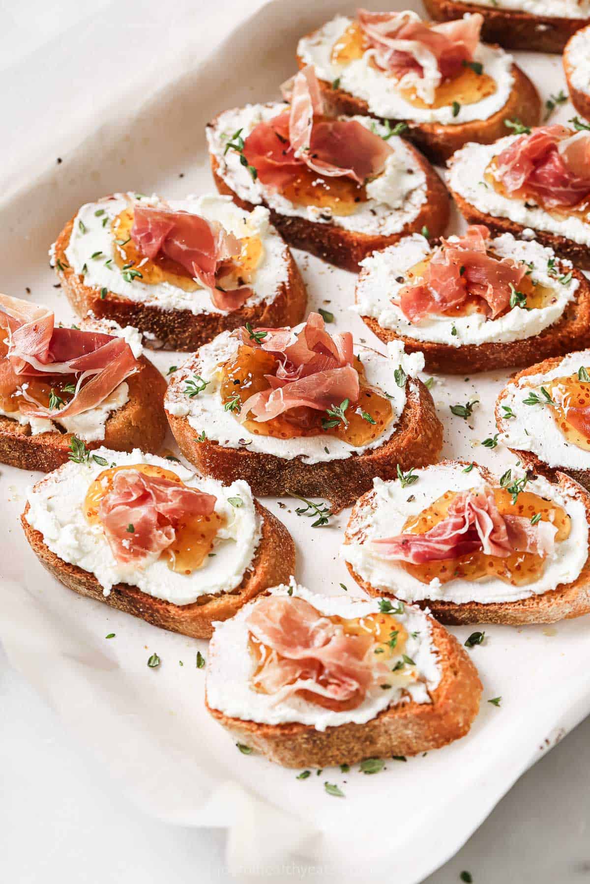 Goat cheese crostinis with fig jam and salty prosciutto on a serving platter.