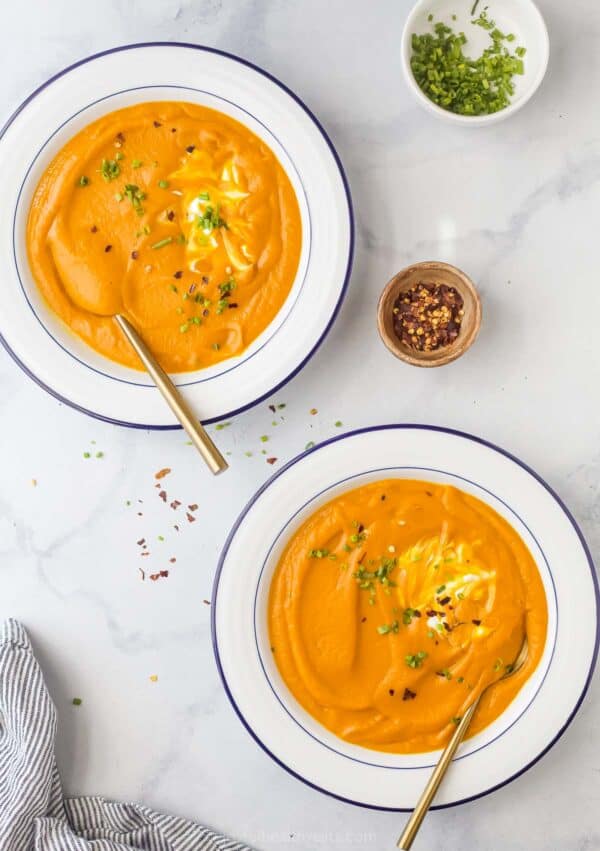 Two bowls of homemade carrot-ginger soup garnished with chives, Greek yogurt, and red pepper flakes.