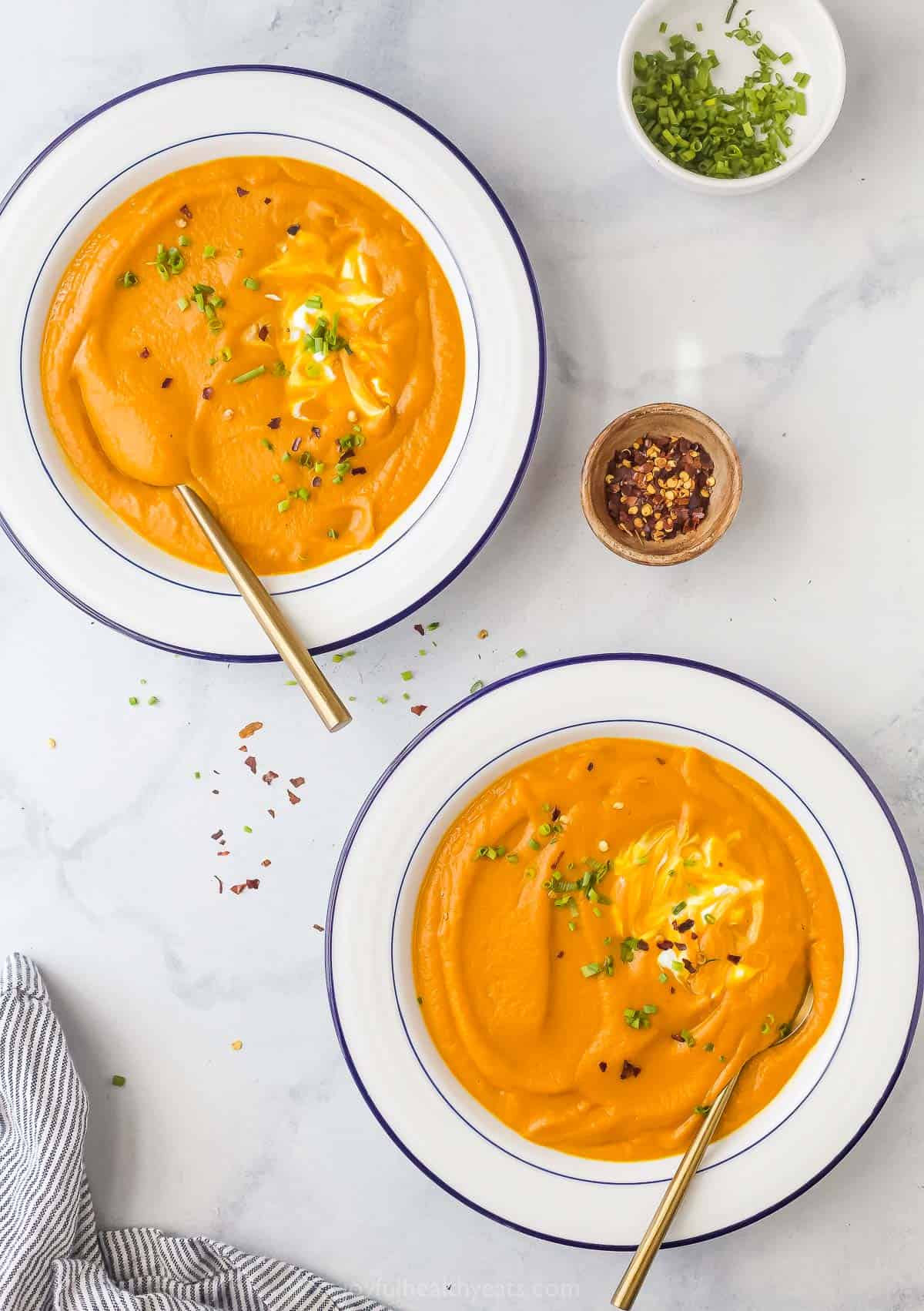 Two bowls of homemade carrot-ginger soup garnished with chives, Greek yogurt, and red pepper flakes. 