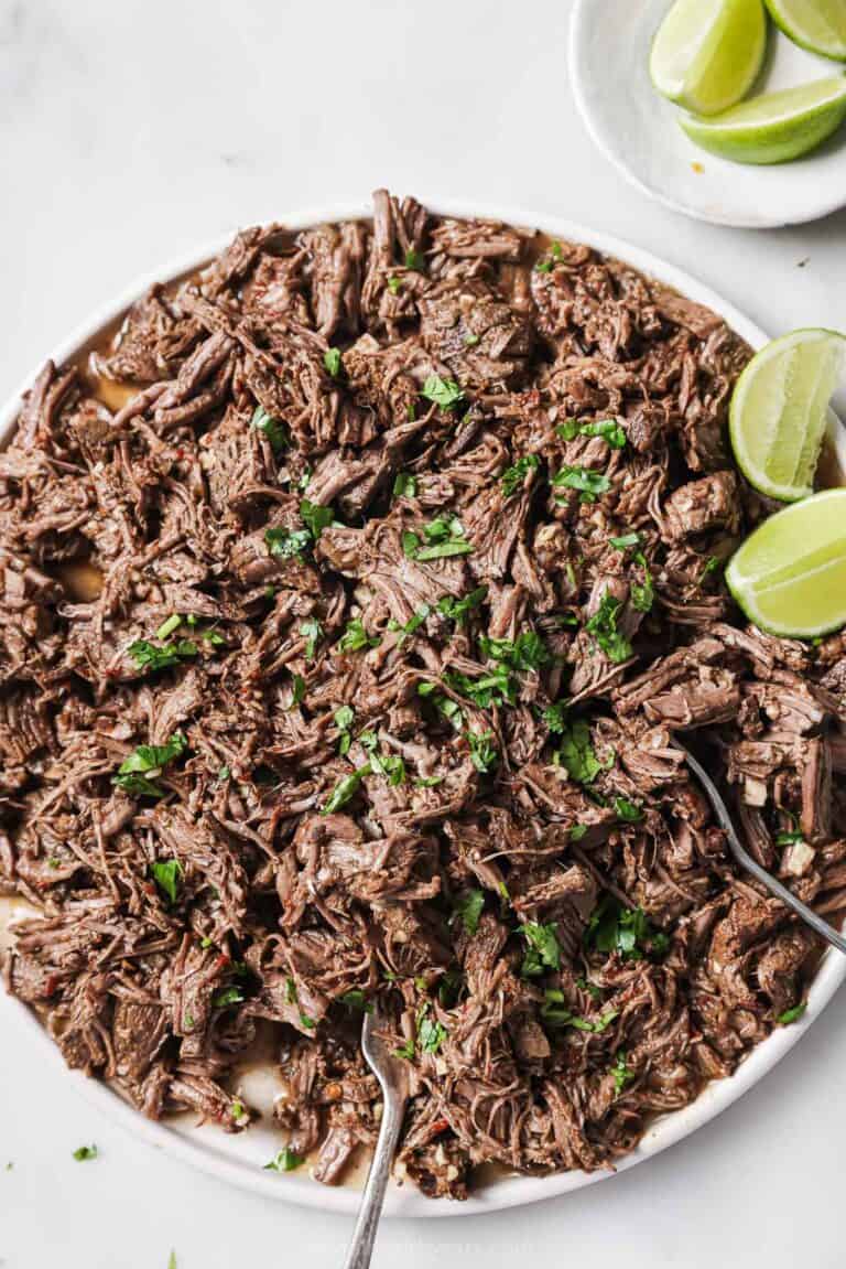 Bowl of chipotle crockpot barbacoa with fresh cilantro with lime wedges on the side.