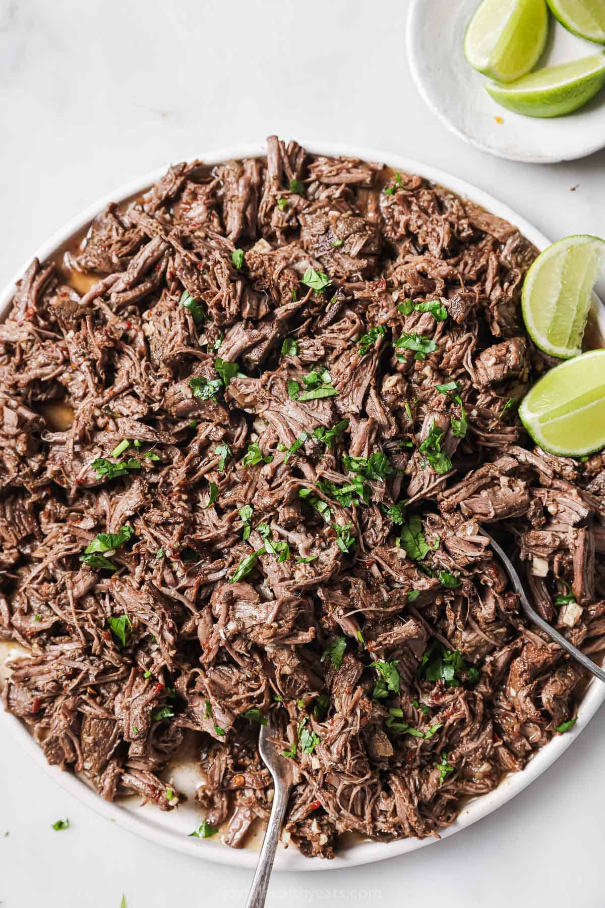 Bowl of chipotle crockpot   barbacoa with fresh cilantro with lime wedges on the side. 