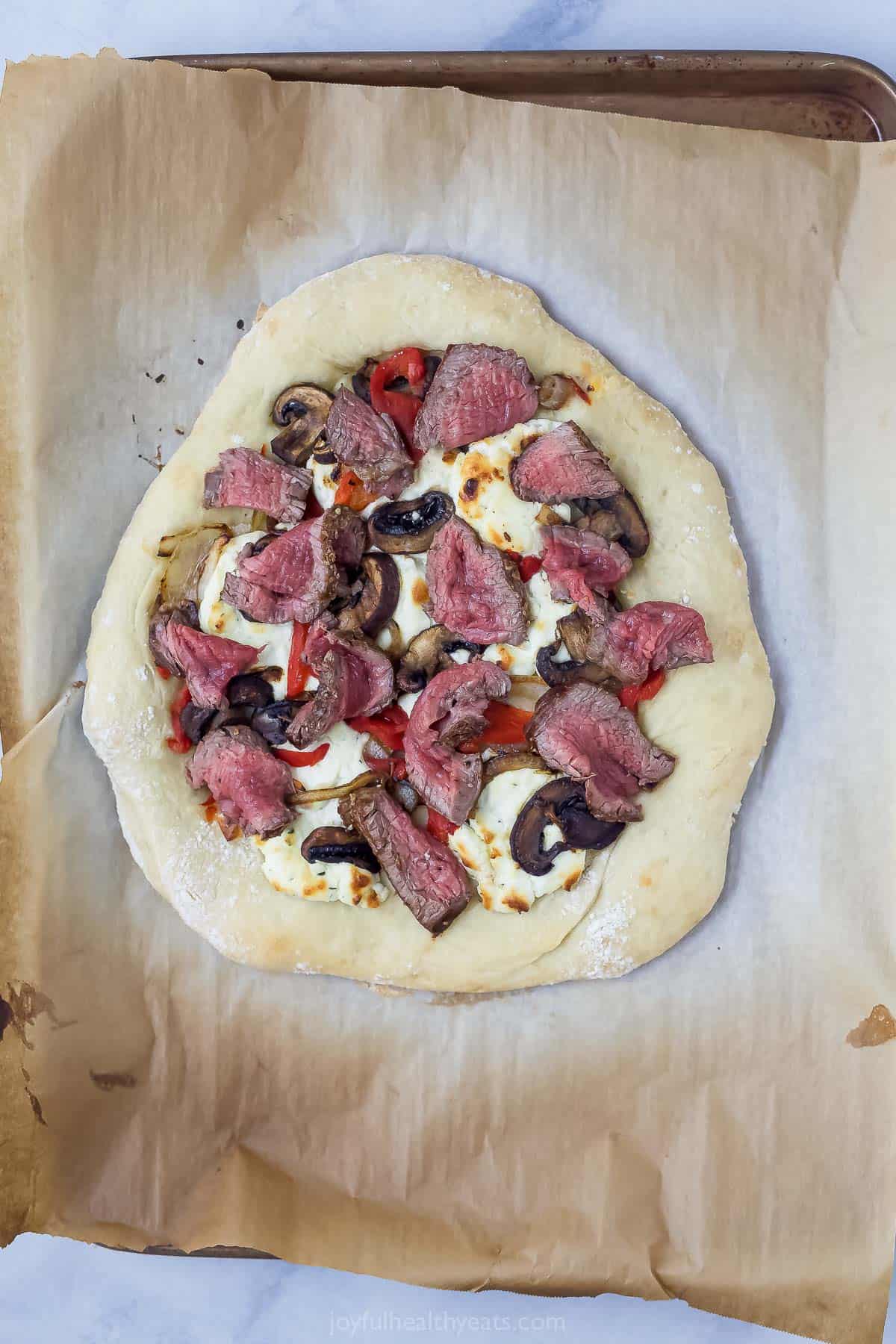 Overhead view of steak pizza on parchment-lined pan before adding arugula