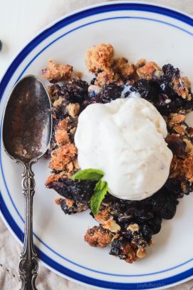 Blueberry crisp on a plate with a scoop of vanilla ice cream and a fresh mint sprig on top.