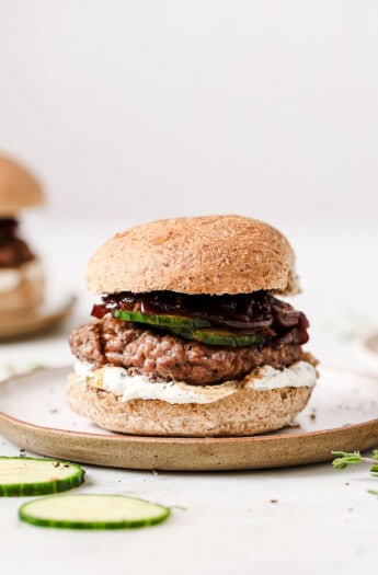 A lamb burger with whipped feta, caramelized onions, and sliced cucumbers on a serving plate.