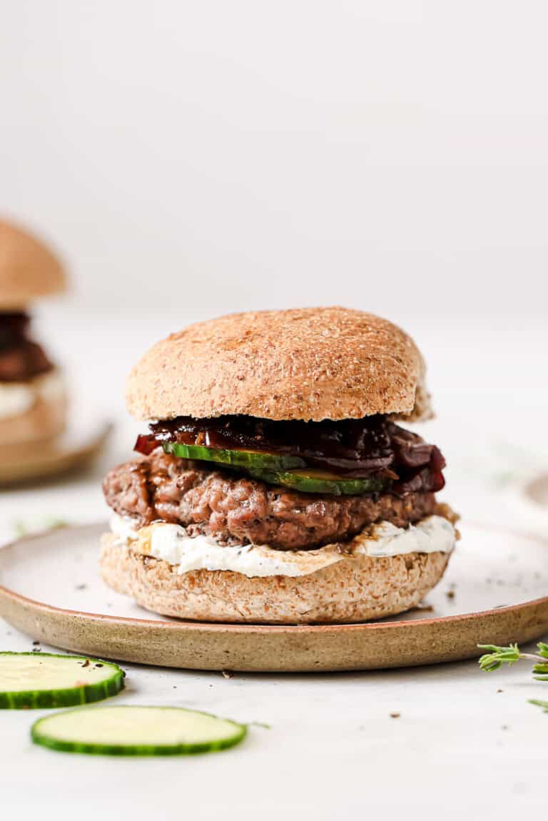 A lamb burger with whipped feta, caramelized onions, and sliced cucumbers on a serving plate.