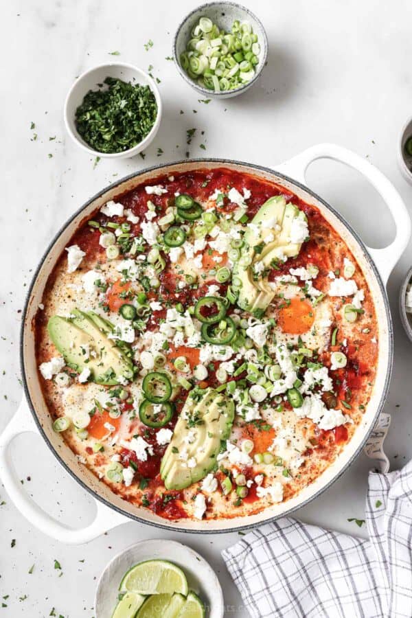 Saucy shakshuka in the skillet with avocado, cilantro, spicy jalapeños, and creamy queso fresco on top.