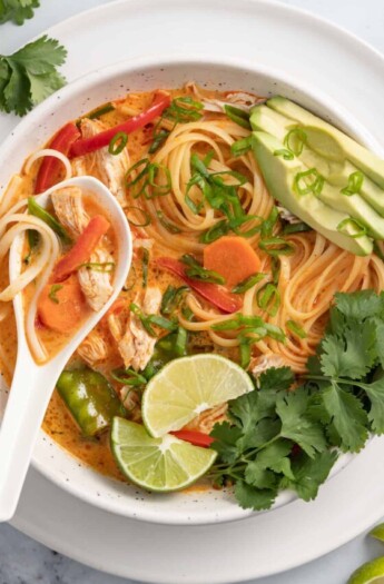 thai chicken noodle soup with an orange colored broth, rice noodles, avocados, and fresh herbs