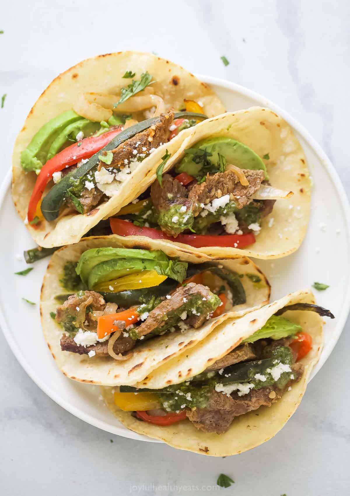 Tacos with beef fajitas, queso fresco, and sliced avocado on a plate. 