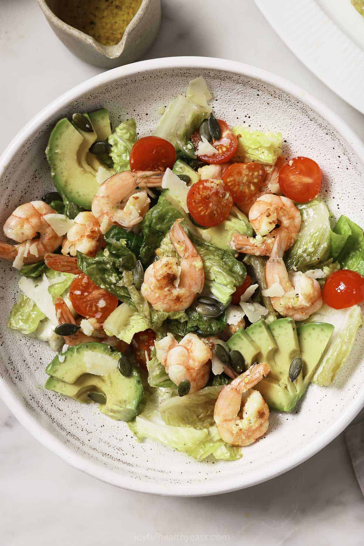 Shrimp caesar salad in a bowl with avocado, tomatoes, lettuce, crunchy pepitas, and dressing. 