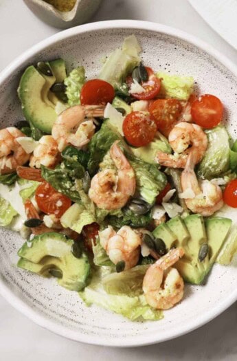 Shrimp caesar salad in a bowl with avocado, tomatoes, lettuce, crunchy pepitas, and dressing.