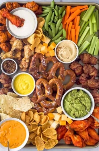 an assortment of snack foods on a tray like pretzels, chicken wings, cheese, veggie sticks, and tortilla chips scattered around dips like guacamole, mustard, and ranch