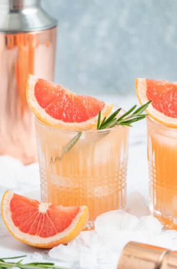 a pale pink beverage known as a greyhiound cocktail in a lowball glass with grapefruit wedge and fresh herb garnish