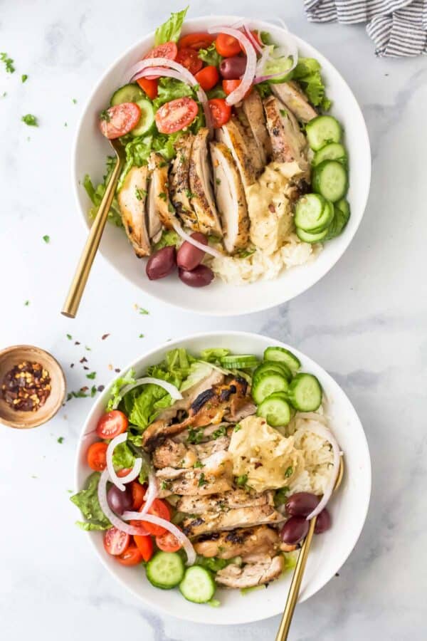 Two Greek chicken bowls with homemade vinaigrette, olives, veggies, rice, and hummus.