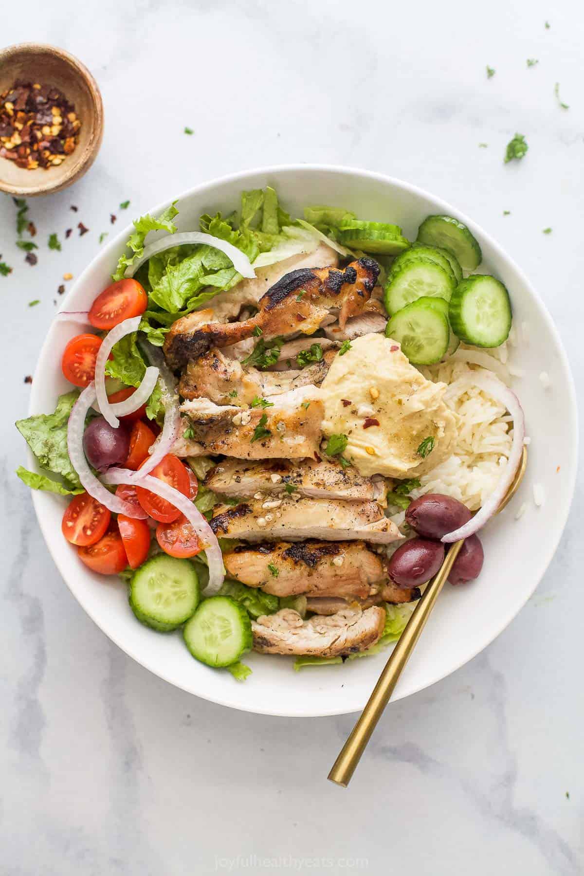 Overhead photo of a grilled chicken bowl with Greek vinaigrette, fresh veggies, olives, and hummus. 